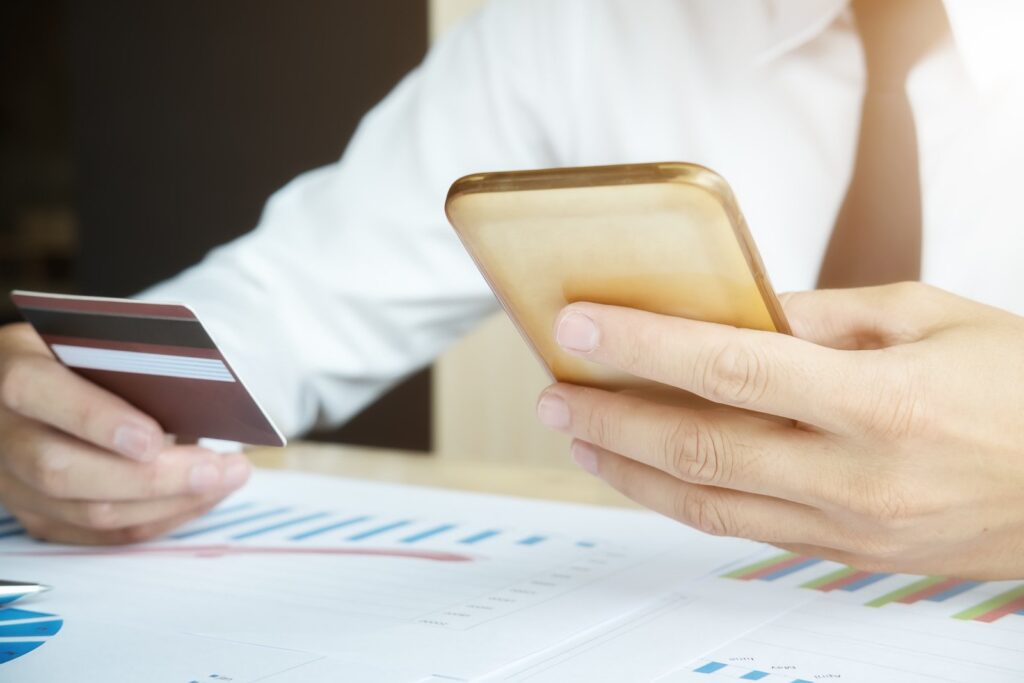 Homem de camisa branca e gravata preta segurando celular dourado e cartão de crédito Depois BBVA Gold, papéis com gráficos sobre a mesa