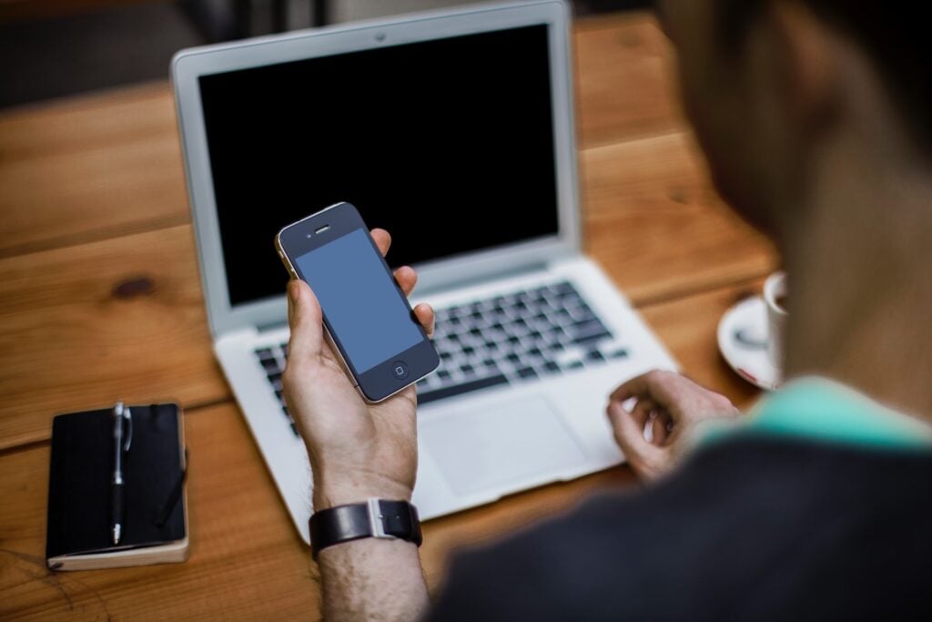 Homem segurando celular, e notebook sobre a mesa