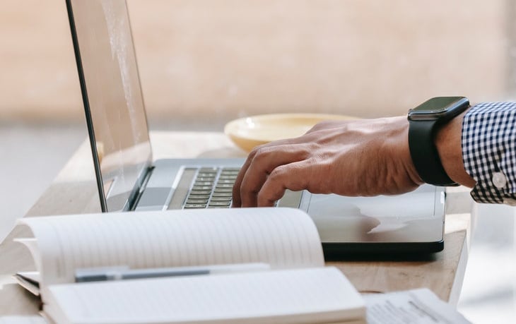 Mão masculina vestindo camisa azul xadrez e relógio, mexendo em notebook sobre mesa, com pires de um lado, e do outro lado um caderno aberto com uma caneta no meio sobre ele