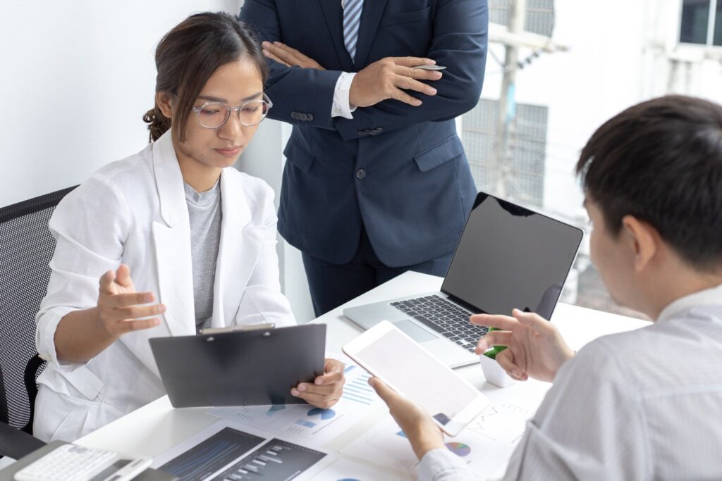 Mulher vestida com blazer branco dando orientações financeiras a dois homens, segurando uma prancheta e sentada em uma cadeira de escritório. Notebook e documentos sobre a mesa do escritório. Um homem está em pé ao lado dela com os braços cruzados e traja um terno, o outro está sentado em frente à mulher e segura um tablet, trajando uma camisa branca social.