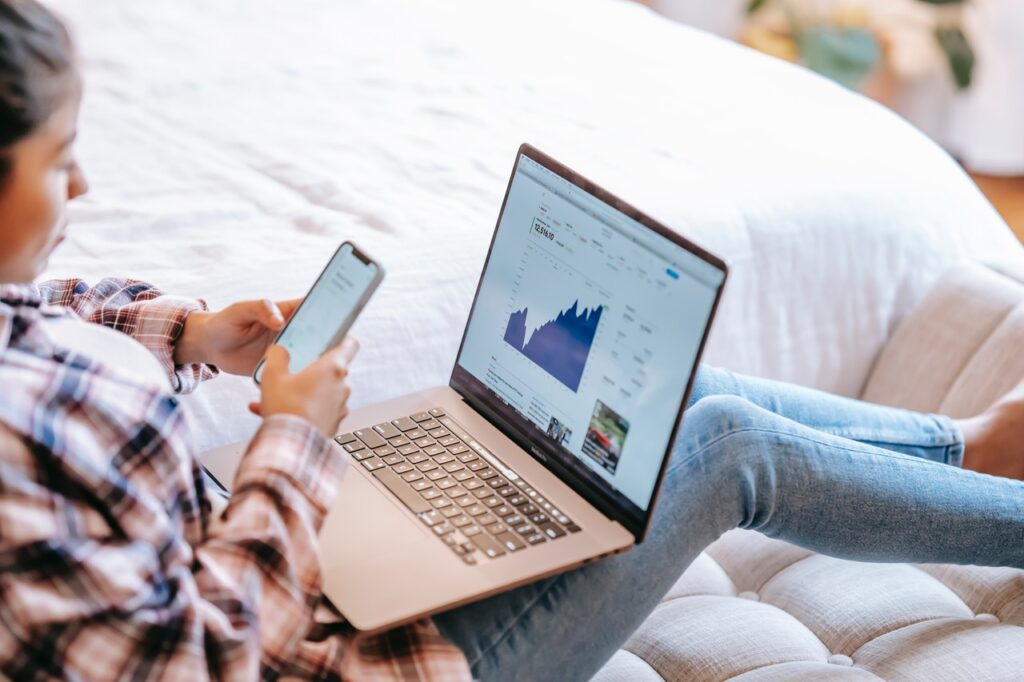 Mulher com notebook no colo e segurando celular. tela do notebook mostra u gráfico crescente, aparentemente lucrativo