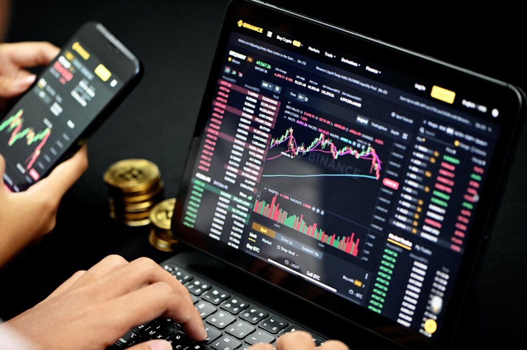Plataforma da corretora Binance em telas de notebook e celular, com gráficos e transações de cripto. Moedas ao fundo representando criptomoedas. Mãos mexendo nos dispositivos.