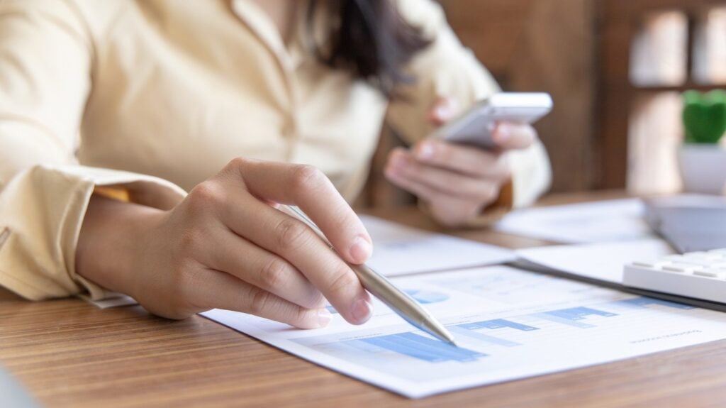 Mulher usando celular e analisando documento com gráfico comparativo