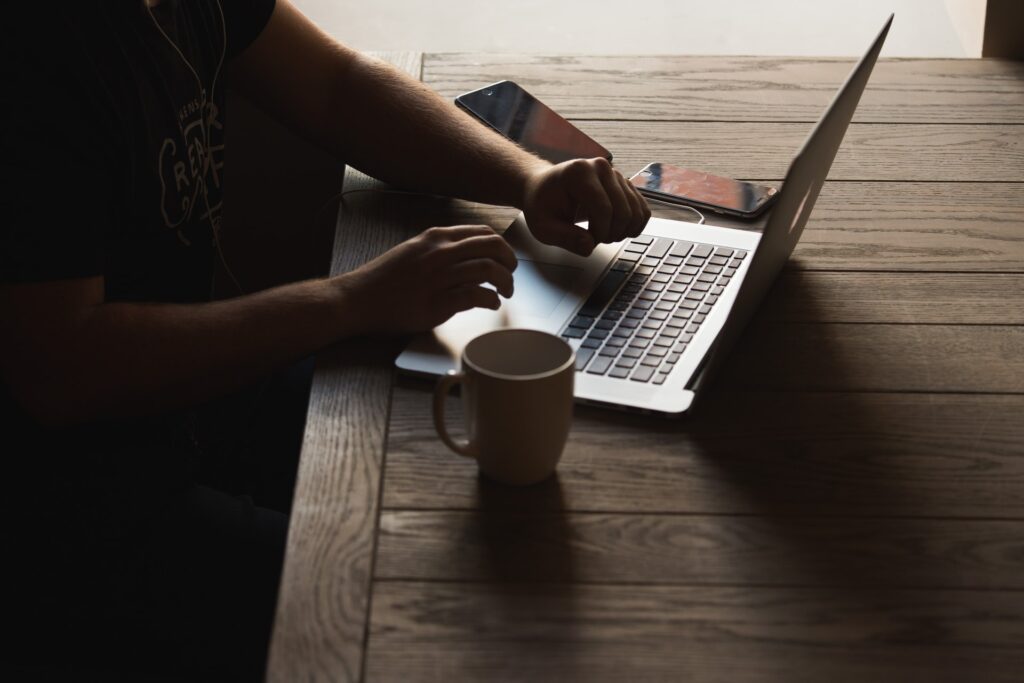 Dois celulares, caneca e notebook sobre mesa de madeira. Pessoa usando o notebook, que pode ser usado para aderir à corretora do Banco Invest.
