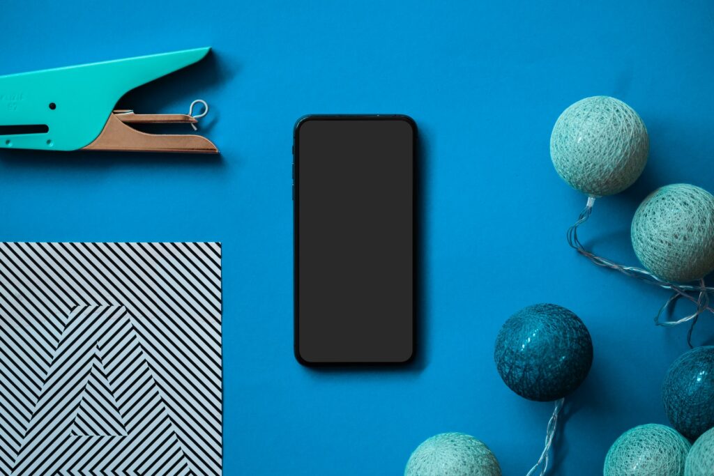 Celular, com o qual se pode aderir à corretora Degiro online, sobre mesa azul (cor da identidade visual Degiro) com enfeites azuis ao redor, grampeador e papel decorativo com padrões diagonais em preto e branco formando a letra A