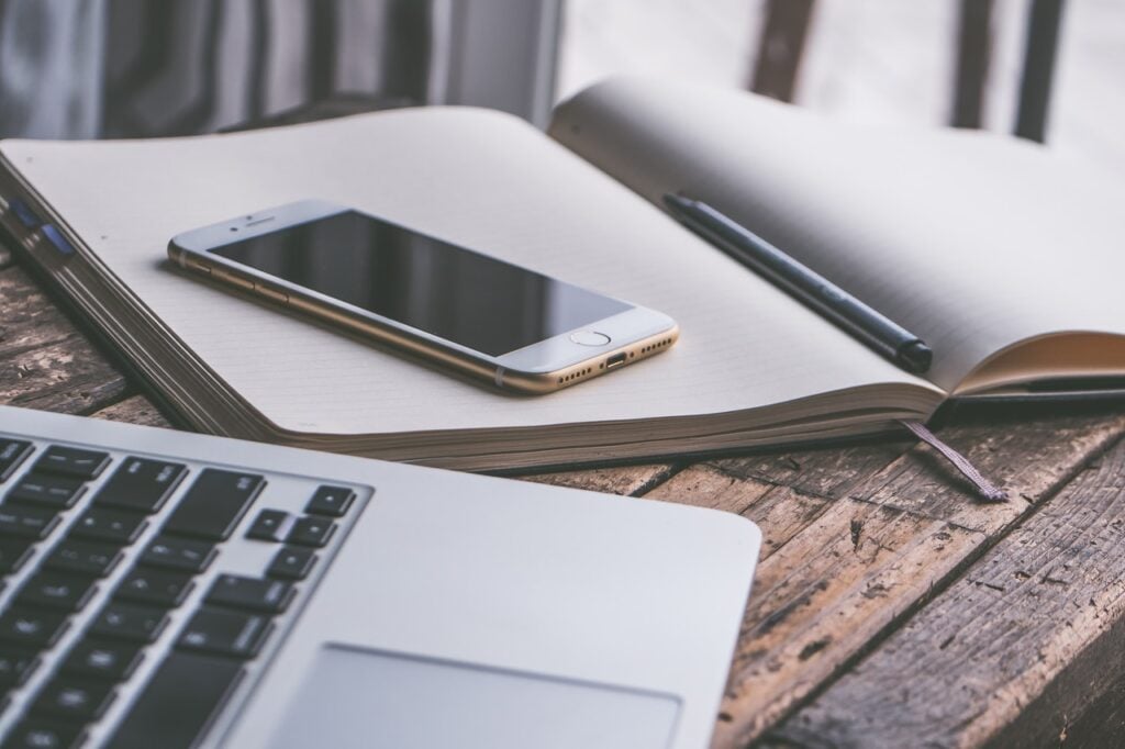 Caderno, caneta, celular e notebook sobre a mesa. Representando como é possível como aderir ao Novo Banco online