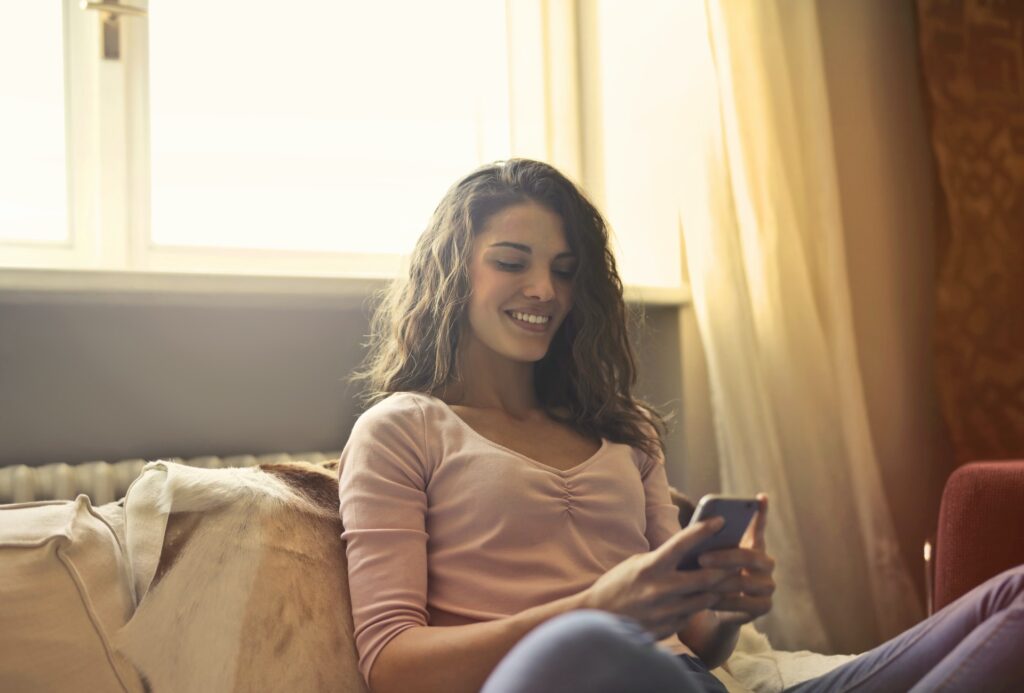 Mulher sentada em sofá usando celular, onde pode-se usar a conta Neteller online. Janela com cortina ao fundo