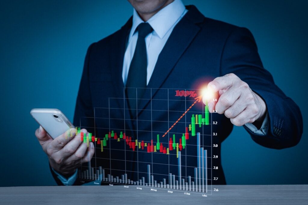 Homem de terno segurando celular com uma mão, e com a outra mão apontando para um gráfico digital de investimento crescente/positivo. Como na corretora Banco Invest, que funciona com investimentos variados e possui aprendizagem online gratuita.