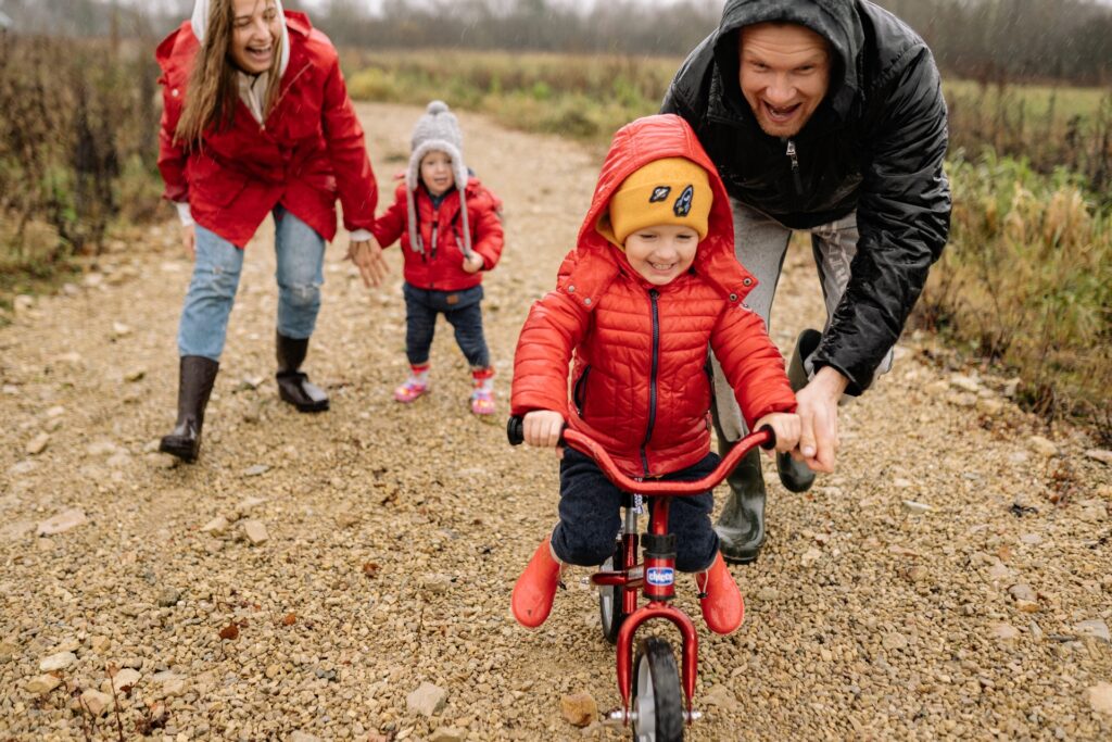 Família se divertindo, simbolizando o Auxílio Filho. Pai ensinando o filho criança a andar de bicicleta, mãe atrás segurando a mão do filho bebê sorrindo observando a cena