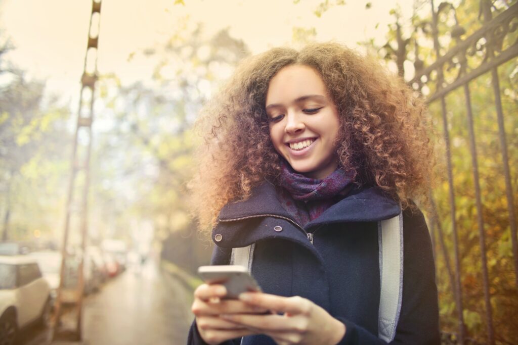Mulher com cabelos cacheados em ambiente externo, sorrindo, segurando e olhando para o celular, com o qual é possível usar Skrill e descobrir o que é