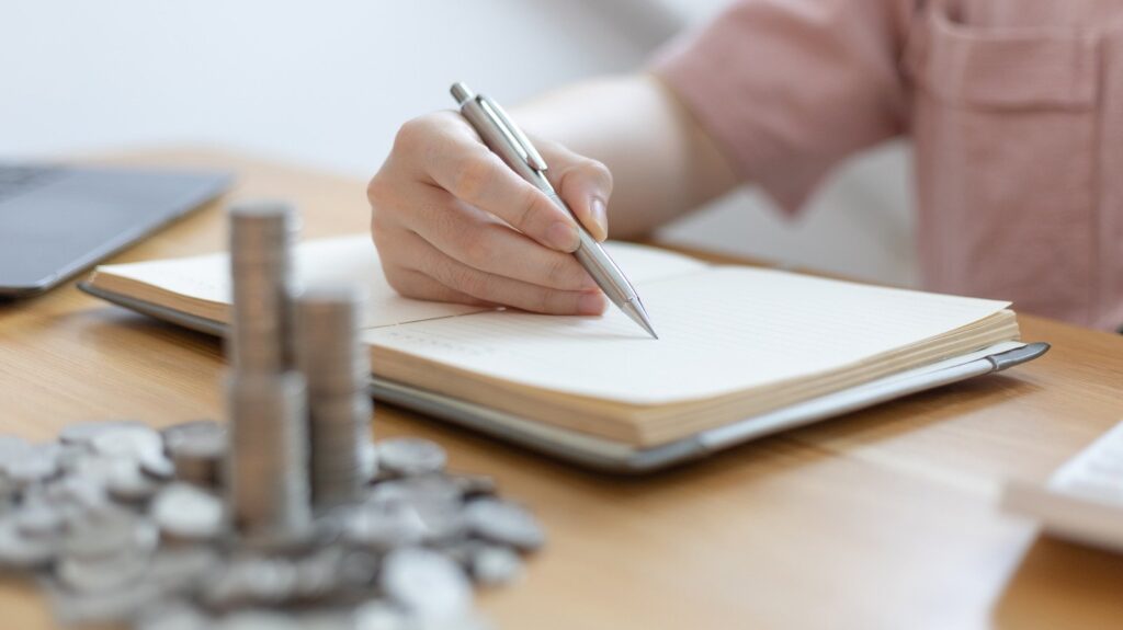 Pessoal antando algo num caderno que está sobre a mesa. Notebook ao fundo. Uma grande quantidade de moedas também sobre a mesa, simbolizando o crédito pessoal BBVA.