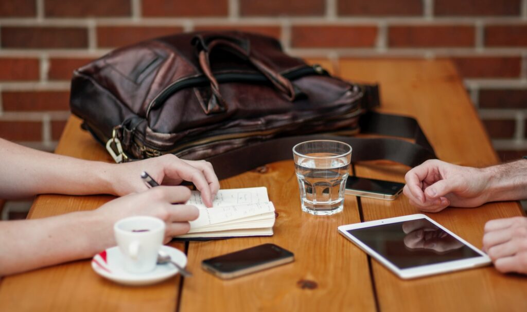 Par de mãos, mochila, copo com água, xícara sobre a mesa. Uma das pessoas está anotando algo num caderninho sobre a mesa. Além disso, também sobre a mesa: celular e tablet, que podem ser usados para solicitar o crédito pessoal Santander Totta