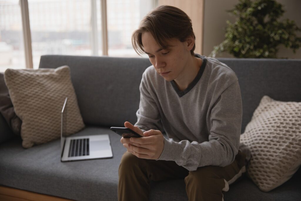 Jovem rapaz segurando celular sentado em sofá. Sobre o sofá também almofadas e um notebook. Representando como solicitar cartão de débito Novo Banco online