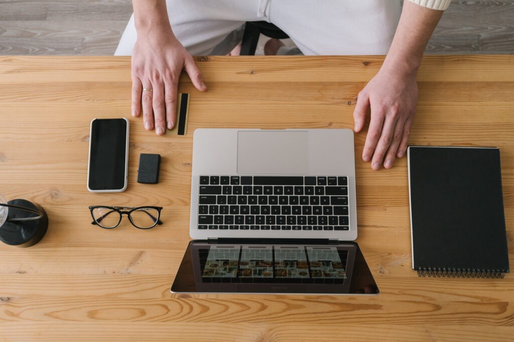 Pessoa sentada à mesa de escritório. Sobre a messa: notebook, tablet, óculos e celular. A mão da pessoa está sobre a mesa, segurando um cartão, que simboliza os cartões BCP