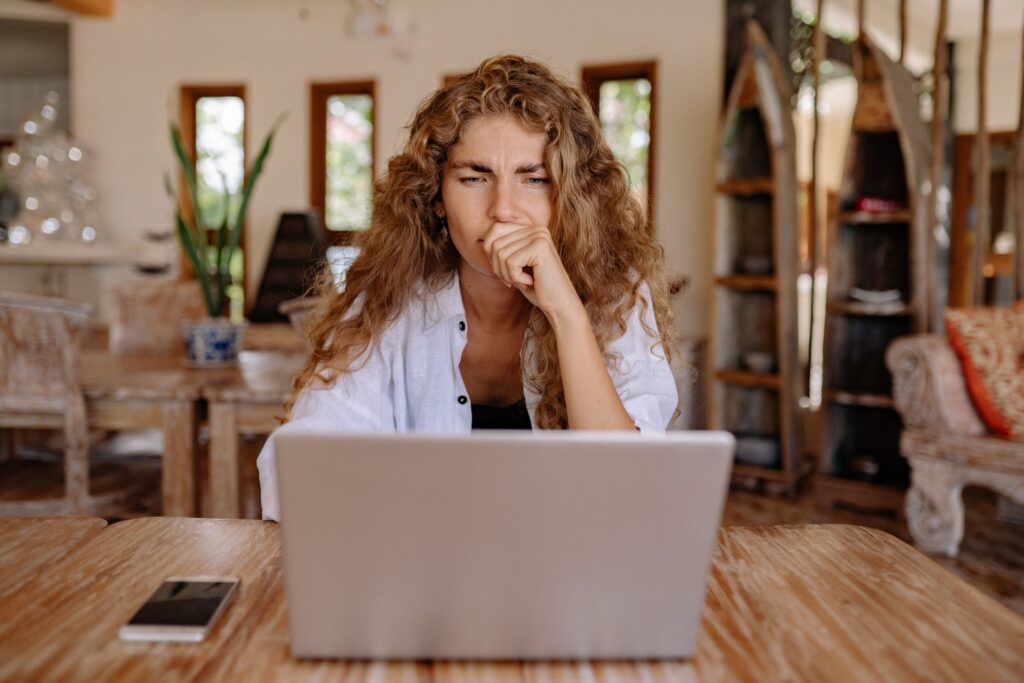 Mulher dentro de casa olhando para a tela de notebook com expressão reflexiva. Também sobre a mesa, ao lado do notebook, um celular.. Simbolizando formas de solicitar o crédito Cetelem online