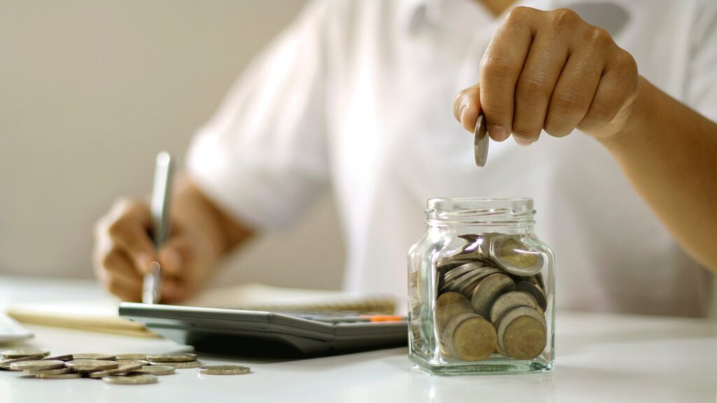Pessoa escrevendo com uma mão, calculadora ao lado do bloco de anotações, e com a outra mão a pessoa está colocando uma moeda num pote cheio de moedas, simbolizando o crédito Pessoal CrediJet