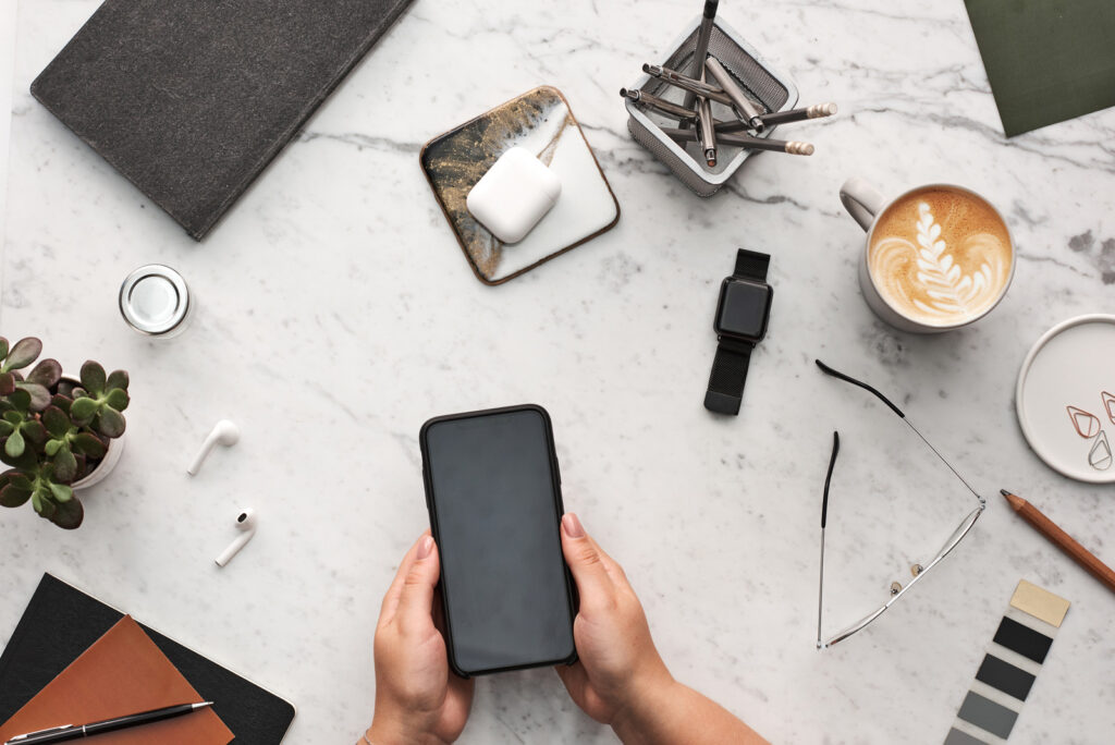 Pessoa usando celular. O fundo é uma mesa de mármore que contém sobre ela: vaso de plantas, fones de ouvido sem fio e carregador, bloco de anotações com caneta, caneca com arte feita com leite, óculos, lápis, clipes e post-its. Simbolizando a diversidade e praticidade do crédito consolidado Gestlifes