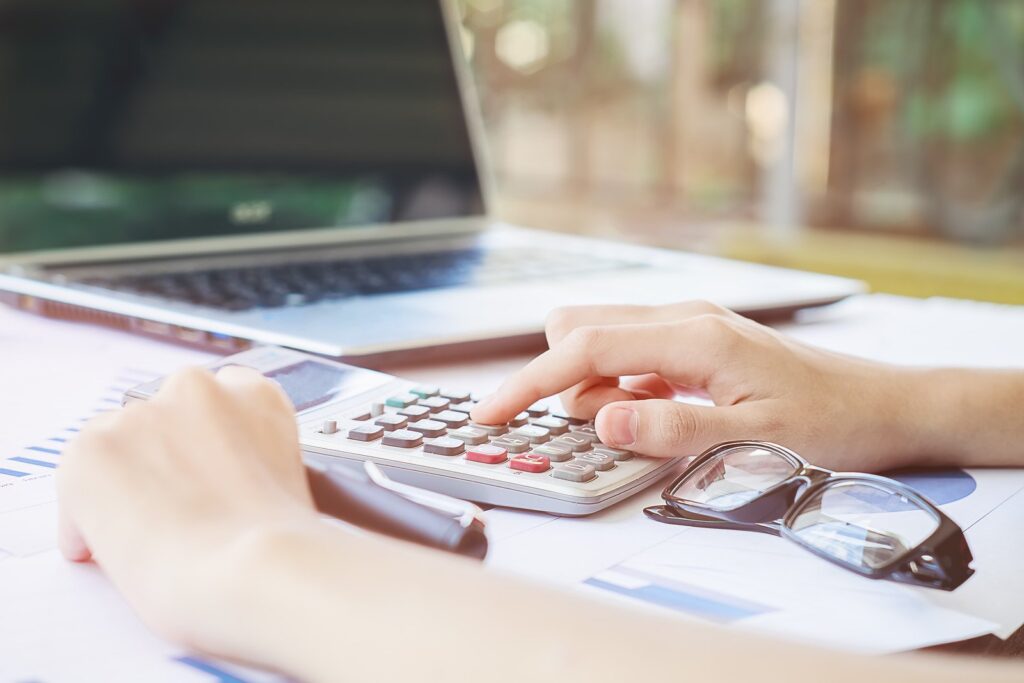 Sobre uma mesa de escritório, documentos, caneta e óculos. E mão mexendo em calculadora que está em frente a um notebook. Simbolizando como solicitar crédito Instacrédito online