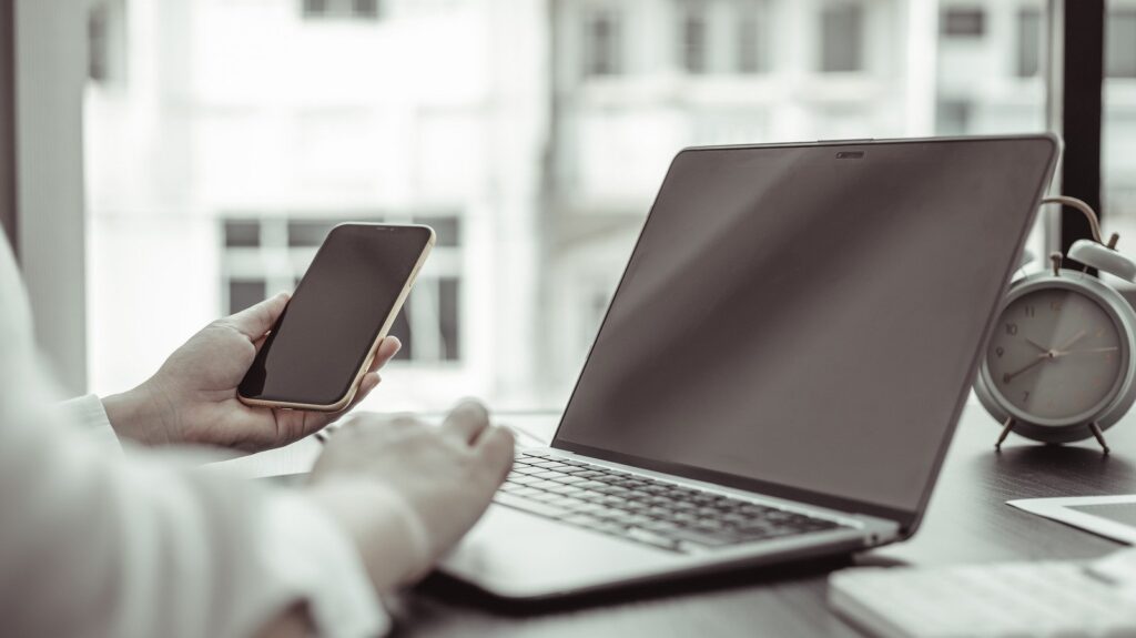 Pessoa à mesa de um escritório, segurando celular enquanto usa notebook. Relógio despertador ao fundo sobre a mesa. Simbolizando como simular e contratar o crédito Montepio
