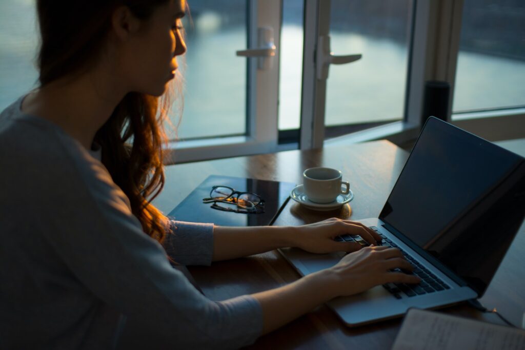Mulher usando notebook. Xícara, óculos e caderno também sobre a mesa. Esse ato de navegar online simbolizando como solicitar o crédito Tenha Menos Dívidas