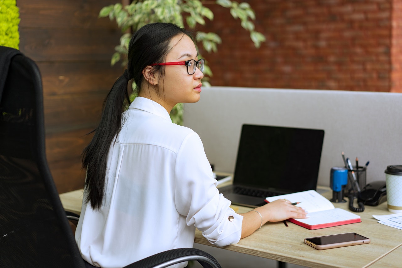 Jovem menina de óculos sentada à mesa de escritório, segurando lápis sobre agenda. Além da agenda, também sobre a mesa: porta canetas com canetas e lápis, carimbo, copo de cafeteria, celular e notebook