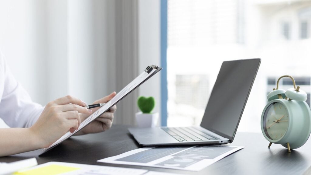 Pessoa à mesa de escritório analisando prancheta com dados. Cobre a mesa estão documentos com gráficos e notebook, e um relógio despertador. Simbolizando opções crédito aprovado na hora.
