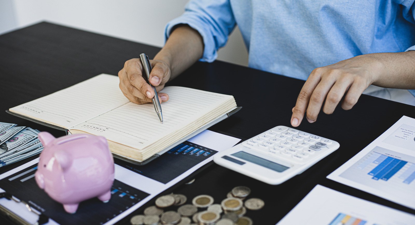 Pessoa escrevendo em agenda sobre mesa de escritório enquanto usa calculadora. Também sobre a mesa estão: documentos com gráficos, dinheiro em cédula e moedas, e um cofrinho de porquinho. Simbolizando opções crédito aprovado na hora.