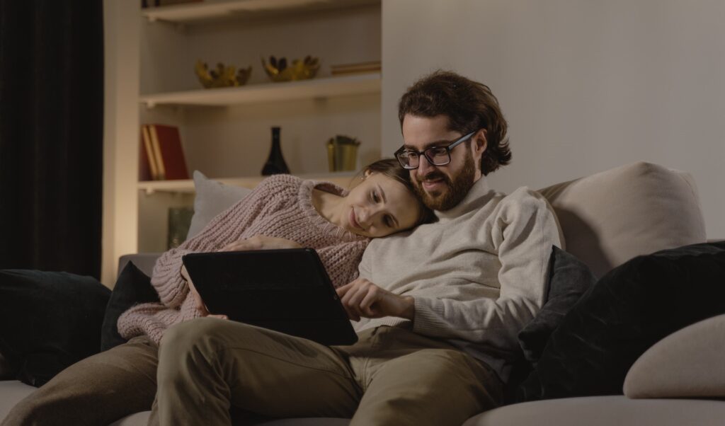 Casal no sofá, mulher com cabeça no ombro do homem, olhando para tela do tablet. E homem segurando o tablet. Representando como acessar o simulador ComparaJá online.