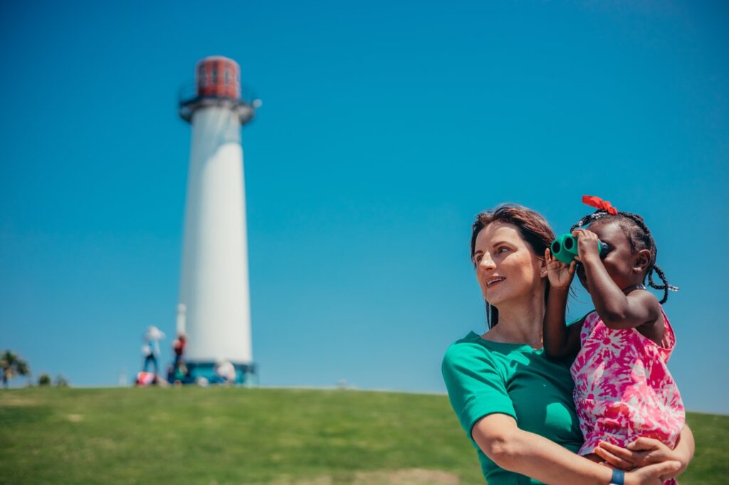 Farol ao fundo, paisagem externa com grama. Em foco uma mulher olhando para o horizonte segurando no colo uma criança de binóculos. Simbolizando o olhar esperançoso para o futuro, que o crédito pessoal Credibom pode oferecer.