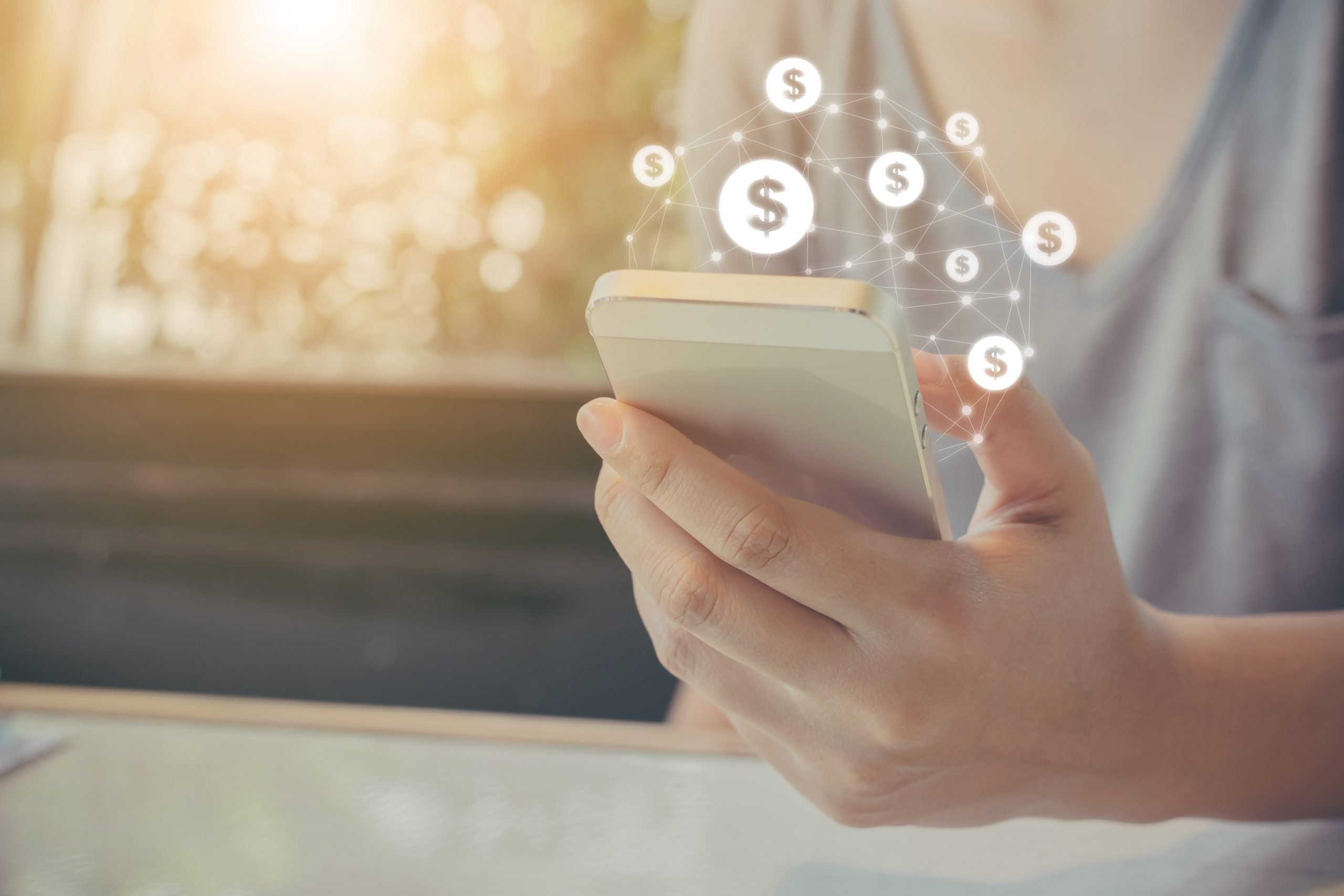 Mão feminina segurando celular com cifrões flutuando sobre a tela. Simbolizando jogos que dão dinheiro no paypal