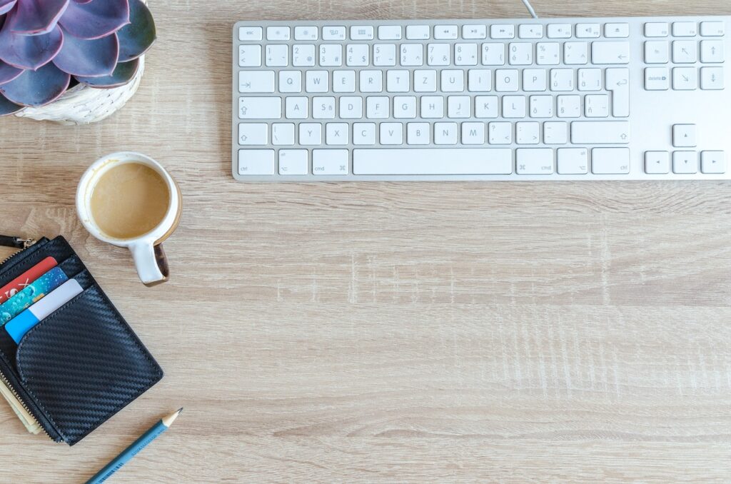 teclado de computador ao lado de vaso de plantas e caneca de café com leite, além de uma carteira com cartões e um lápis