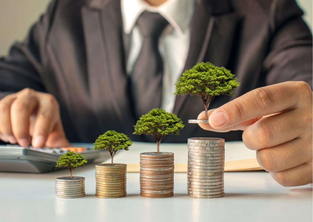 pessoa de terno e gravata segurando árvore em miniatura e empilhando moedas em cima da mesa