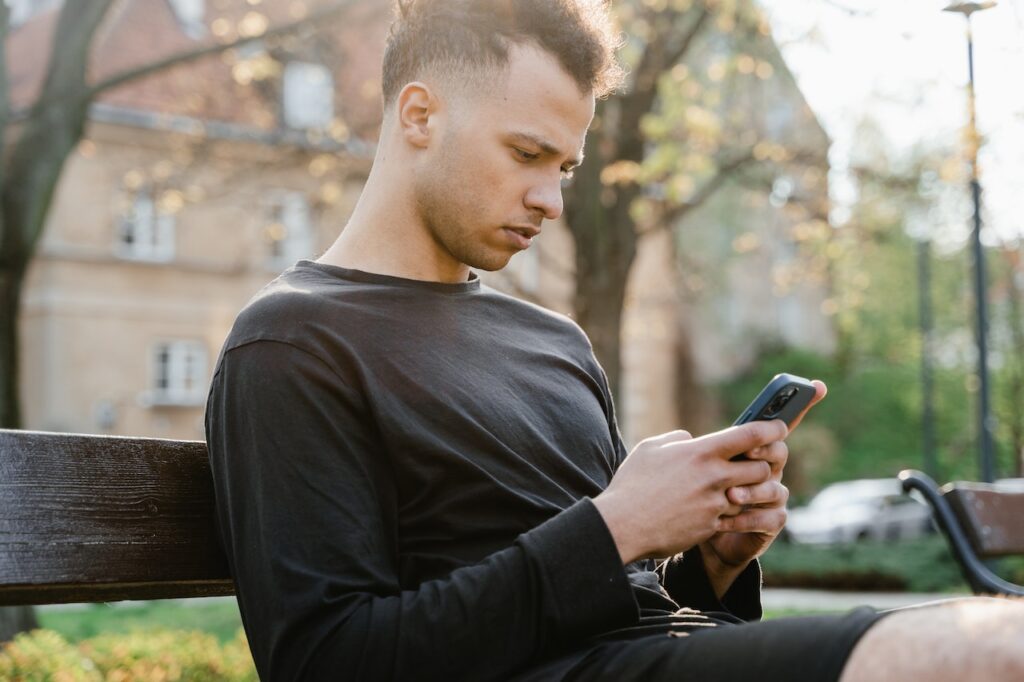 homem sentado em banco de praça mexendo no celular