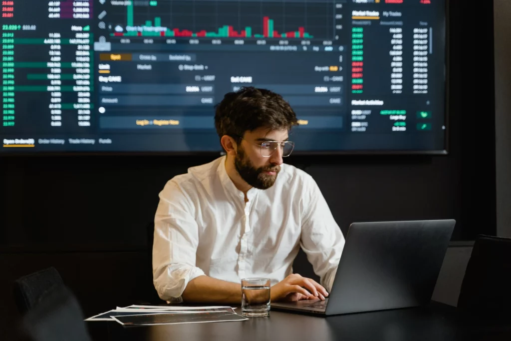 Homem concentrado olhando notebook com painel de investimentos ao fundo