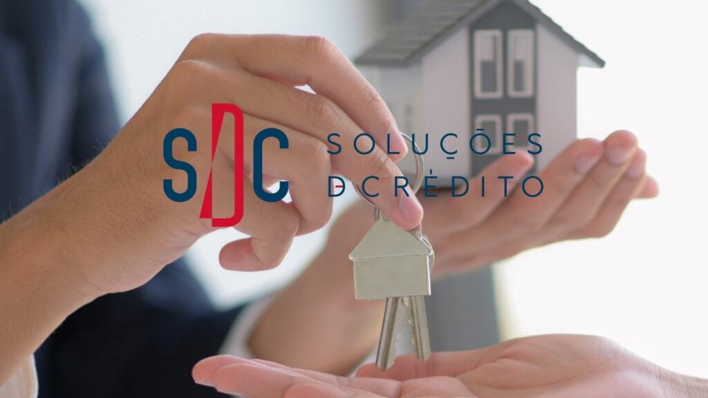 Mão entregando chave com miniatura de casa ao fundo