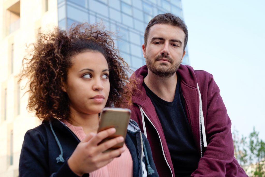 Mulher desconfiada usando celular e homem atrás espiando por cima do ombro