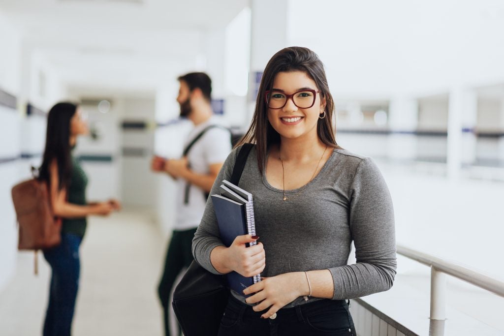 Portrait of young Brazilian student with backpack carrying books in college. Other students in the background.