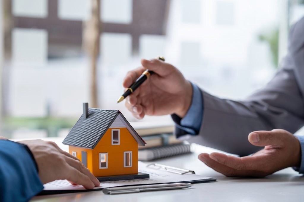 Mão segurando caneta exlicando contrato e miniatura de casa ao lado