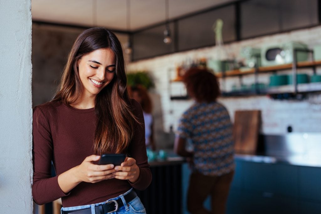Young woman smiling while using phone while standing in kitchen